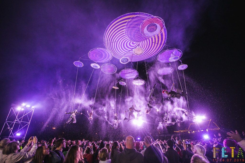 zdjęcie; konstrukcja z kilkunastu kół różnej wielkości na tle nocnego nieba, pod konstrukcją na linach podwieszonych jest kilkunastu tancerzy powietrznych, rzucają konfetti na publiczność, oświetleni na fioletowo. Pod nimi duża publiczność.